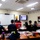 A classroom training session is taking place at the MCIPAC F&amp;ESJ Fire Academy, where an instructor stands at a red podium delivering a presentation to a group of firefighters. Students from various fire departments, including U.S. Army Okinawa Fire Department and Chatan Fire Department, are seated at tables with textbooks and materials. A presentation is displayed on a wall-mounted monitor behind the instructor.