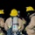 Three firefighters in full turnout gear are gathered together in what appears to be a nighttime or low-light environment, possibly during a training exercise or active incident. All personnel are equipped with SCBA (Self-Contained Breathing Apparatus) tanks and yellow hard hats. One firefighter facing the camera wears a mask and a cap reading '03 OKINAWA,' suggesting a U.S. military or MCIPAC (Marine Corps Installations Pacific) fire department affiliation.