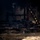 The image shows the interior of a structure following a significant fire event. Charred and collapsed wooden debris, likely the remains of furniture or structural elements, is visible on a wet floor covered in ash and soot. A firefighter's gloved hand is partially visible in the upper right corner, suggesting active post-fire assessment or overhaul operations.