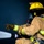A firefighter in full personal protective equipment is actively operating a fire hose, directing a high-pressure water stream toward an unseen target. The firefighter is wearing standard turnout gear including a yellow helmet, SCBA (Self-Contained Breathing Apparatus) with a Scott Air-Pak tank, and reflective striped bunker coat. A second firefighter is partially visible in the background, also wearing a yellow helmet.
