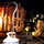 Two firefighters in full turnout gear and SCBA (Self-Contained Breathing Apparatus) are crouched low near a burning wooden pallet structure during what appears to be a controlled fire training exercise. A propane tank with an attached hose and hand truck is visible in the foreground, suggesting the fire is deliberately fueled for training purposes. The scene is set in a dark environment, likely an indoor or enclosed training facility.