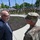 Acting SecAF Roth visits Joint Base Andrews A civilian in a blue suit is engaged in conversation with a military officer in OCP (Operational Camouflage Pattern) uniform in the foreground. Behind them, a large formation of soldiers in military uniforms stands at attention in an outdoor setting near a government or military building. The scene appears to be a formal military ceremony or official visit.