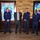 Acting SecAF Roth visits Joint Base Andrews Six individuals pose for a formal group photo in what appears to be a military venue or club. Five of the individuals are wearing matching dark navy blue uniform jackets with insignia patches, while one person in the center is dressed in a civilian business suit. Two decorative stained glass windows featuring 'Sam Fox' squadron artwork with military aircraft motifs are visible in the background.