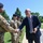 Acting SecAF Roth visits Joint Base Andrews A civilian man in a dark suit is shaking hands with a female military service member in multicam/OCP uniform and black beret, while additional uniformed personnel stand in a line behind her. The setting appears to be an outdoor military installation or base on a clear, sunny day. The interaction appears ceremonial or formal in nature, suggesting a visit or inspection.
