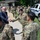 Acting SecAF Roth visits Joint Base Andrews A civilian official in a suit is shaking hands with U.S. Air Force Security Forces (SF) personnel in military camouflage uniforms during an outdoor meet-and-greet event. A group of uniformed airmen stands in formation in the background near a brick building. A white police vehicle marked 'POLICE' and 'DIAL 9-1-1' is visible on the right side of the image.