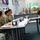 Acting SecAF Roth visits Joint Base Andrews A civilian man in a dark suit is seated across from a group of military personnel in camouflage uniforms at a dining facility or conference setting. The gathering appears to be a formal meeting or briefing between the civilian official and service members. Name placards are visible on the tables, suggesting a structured, organized engagement.