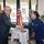 Acting SecAF Roth visits Joint Base Andrews A civilian man in a blue pinstripe suit is shaking hands with a female U.S. Air Force service member in a dark blue uniform. The ceremony appears to take place in a building lobby or reception area, with an American flag and a POW/MIA flag visible in the background. A small ceremonial table with a book (likely a Bible) and other items is positioned between them.
