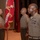 Three U.S. Marine Corps personnel are participating in a formal military ceremony, likely a change of command or similar official event. One Marine on the left is holding a red unit guidon/colors with gold fringe, while two senior Marines stand at attention facing him. The setting appears to be an auditorium or ceremonial hall with multiple flags displayed in the background.