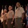 Three U.S. Marine Corps officers in service uniform (tan/khaki shirts with olive green trousers) stand at attention on a stage, looking upward, likely during a formal military ceremony. They are positioned in front of a backdrop of multiple U.S. state flags arranged on staffs against a black curtain. The officers display various service ribbons and medals on their uniforms, indicating senior rank and experience.