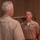 Two military personnel in tan/khaki uniforms are engaged in a face-to-face interaction, likely a formal or semi-formal military exchange. The officer facing the camera displays multiple service ribbons, a naval aviator or similar warfare device, and rank insignia on his collar. The setting appears to be an indoor military facility or auditorium.