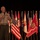 A U.S. Marine Corps officer in service uniform is speaking at a formal event, holding a microphone on what appears to be a stage. He is standing in front of a display of multiple flags including the U.S. national flag, state flags, and a Marine Corps Base flag. His uniform displays multiple service ribbons and medals indicating senior rank.