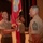 A formal U.S. Marine Corps military ceremony is taking place on a stage or auditorium floor. Three Marines in Service Charlie (tan short-sleeve) uniforms are participating, with one holding a unit guidon or colors flag. The Marine on the right displays Sergeant Major rank insignia (chevrons with a star), indicating a senior enlisted change of command or relief ceremony.