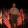 A U.S. Marine Corps officer in service uniform (tan/khaki) stands at a microphone on a stage, appearing to deliver a speech or address. He is positioned in front of a display of numerous U.S. state flags and what appears to be a Marine Corps Base flag. The setting suggests a formal military ceremony or official event.