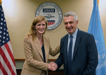 Two individuals are shaking hands in front of a USAID (United States Agency for International Development) seal, flanked by an American flag on the left and a United Nations flag on the right. The setting appears to be an official meeting room, suggesting a formal diplomatic or institutional engagement between USAID and a UN-affiliated organization. Both individuals are smiling, indicating a positive and cooperative interaction.
