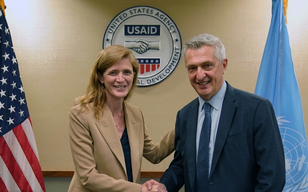 Two individuals are shaking hands in front of a USAID (United States Agency for International Development) seal, flanked by an American flag on the left and a United Nations flag on the right. The setting appears to be an official meeting room, suggesting a formal diplomatic or institutional engagement between USAID and a UN-affiliated organization. Both individuals are smiling, indicating a positive and cooperative interaction.
