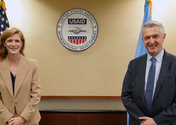 Two individuals are posing for a formal photograph at USAID (United States Agency for International Development) headquarters. They are standing in front of the official USAID seal mounted on a wall, flanked by the American flag and a light blue flag. The setting suggests an official diplomatic or institutional meeting.