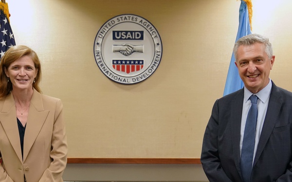 Two individuals are posing for a formal photograph at USAID (United States Agency for International Development) headquarters. They are standing in front of the official USAID seal mounted on a wall, flanked by the American flag and a light blue flag. The setting suggests an official diplomatic or institutional meeting.