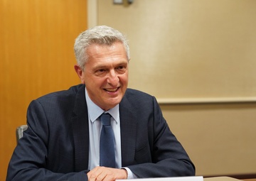 A man is seated at a conference table during what appears to be an official USAID meeting. A nameplate in front of him reads 'HE Filippo Achille Maria Grandi, United Nations High Commissioner for Refugees.' He is smiling and appears engaged in conversation.
