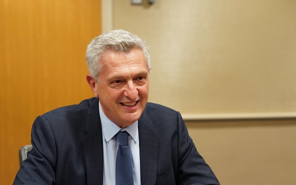 A man is seated at a conference table during what appears to be an official USAID meeting. A nameplate in front of him reads 'HE Filippo Achille Maria Grandi, United Nations High Commissioner for Refugees.' He is smiling and appears engaged in conversation.