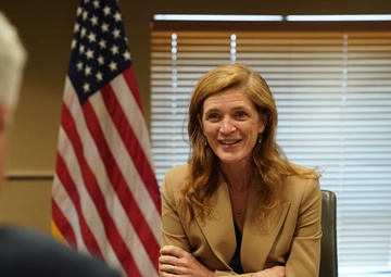 A woman is seated at a conference table in an official government setting, smiling during what appears to be a formal meeting. A nameplate in front of her reads 'Samantha Power, Administrator, USAID'. The setting includes an American flag and a USAID organizational flag in the background.