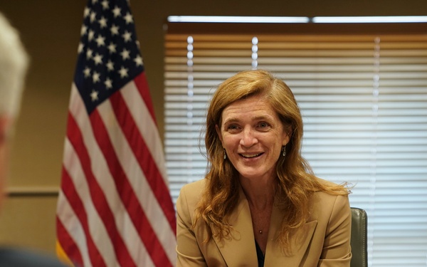 A woman is seated at a conference table in an official government setting, smiling during what appears to be a formal meeting. A nameplate in front of her reads 'Samantha Power, Administrator, USAID'. The setting includes an American flag and a USAID organizational flag in the background.