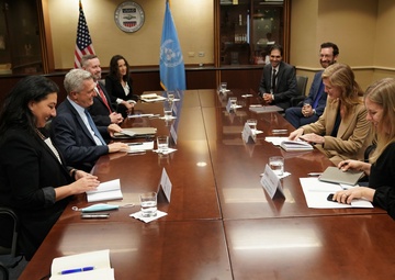 A formal diplomatic or governmental meeting is taking place in a conference room, with two groups of people seated on opposite sides of a large wooden table. The presence of a United Nations flag and a USAID seal/logo on the wall, along with two American flags, suggests this is an official bilateral or interagency meeting. Participants have name placards, documents, and water glasses in front of them.