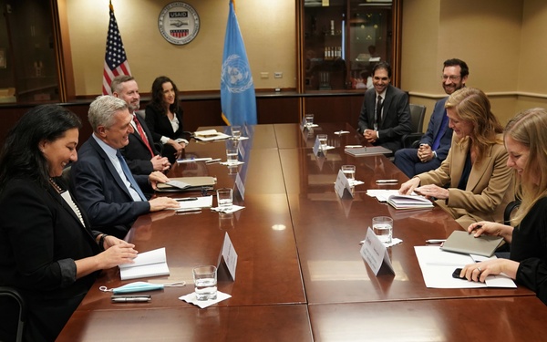 A formal diplomatic or governmental meeting is taking place in a conference room, with two groups of people seated on opposite sides of a large wooden table. The presence of a United Nations flag and a USAID seal/logo on the wall, along with two American flags, suggests this is an official bilateral or interagency meeting. Participants have name placards, documents, and water glasses in front of them.