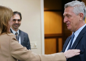 Three individuals are engaged in a professional greeting or meeting in what appears to be an office or conference setting. A woman in a beige blazer is reaching out to touch the arm of an older man in a dark navy suit and blue tie, suggesting a warm professional exchange. A second man in a dark suit with a tie badge is visible in the background, smiling.