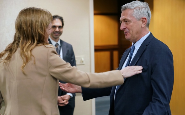 Three individuals are engaged in a professional greeting or meeting in what appears to be an office or conference setting. A woman in a beige blazer is reaching out to touch the arm of an older man in a dark navy suit and blue tie, suggesting a warm professional exchange. A second man in a dark suit with a tie badge is visible in the background, smiling.