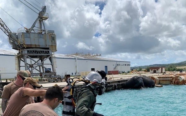 A fully equipped military diver in a hard-hat diving suit is stepping off a pier into turquoise harbor water, assisted by two support personnel on the dock. The scene takes place at what appears to be a naval facility, with a large industrial crane, warehouse buildings, and partially submerged or moored objects visible in the background. One support crew member's shirt reads 'Underwater Construction Team' and 'U.S. Pacific Fleet.'