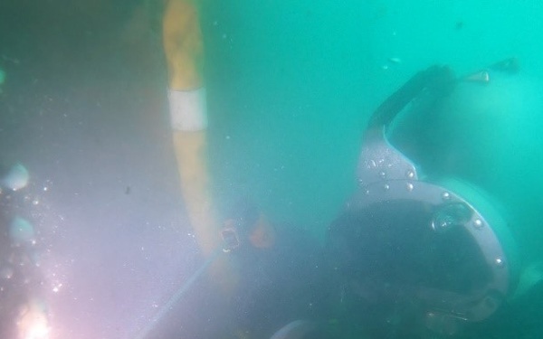 An underwater scene showing a diver in a dark wetsuit performing what appears to be underwater welding or cutting, evidenced by a bright arc light visible on the left side of the frame. The water has a greenish-teal tint with limited visibility due to suspended particles and the intense light source. Bubbles are visible rising from the diver's equipment.