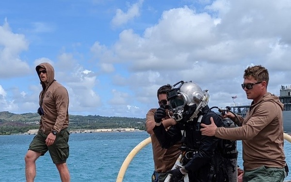 The image shows what appears to be a military diving operation at a naval pier or dock facility. Two personnel in tan/brown military-style clothing are assisting a diver in full hard-hat diving gear, including a metal helmet and dry suit with red boots. A large naval vessel is visible in the background, suggesting this is a military harbor or base.