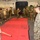 A group of U.S. Marines are conducting a martial arts or close-combat training session inside what appears to be a ship's hangar bay or cargo deck. Personnel are gathered around a red training mat while one instructor demonstrates a technique with a wooden training stick (pugil-style or escrima stick). The setting includes military equipment, crates, and vehicles visible in the background.