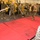 Military personnel, appearing to be U.S. Marines based on their distinctive MARPAT camouflage uniforms, are conducting a martial arts or close-combat training session in what appears to be a vehicle bay or hangar deck. Several participants are holding wooden training sticks (pugil-style or escrima sticks) in fighting stances on and around red training mats. An observer in the foreground watches the demonstration while others stand in a semicircle observing.