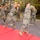 A group of military personnel in MARPAT camouflage uniforms are conducting martial arts or close-combat training with wooden sticks/pugil staves aboard what appears to be a naval vessel's well deck or hangar bay. The trainees are in wide stances performing synchronized striking movements. Red training mats are laid out on the deck floor, and military vehicles and equipment are visible in the background.