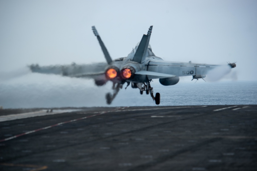 A carrier-based military jet aircraft, marked '500', is captured at the moment of catapult launch from an aircraft carrier flight deck over open ocean. Both afterburners are visibly ignited, producing bright orange flame, and a vapor cone or steam cloud trails behind the aircraft from the catapult steam system. The landing gear is still extended as the aircraft clears the deck.