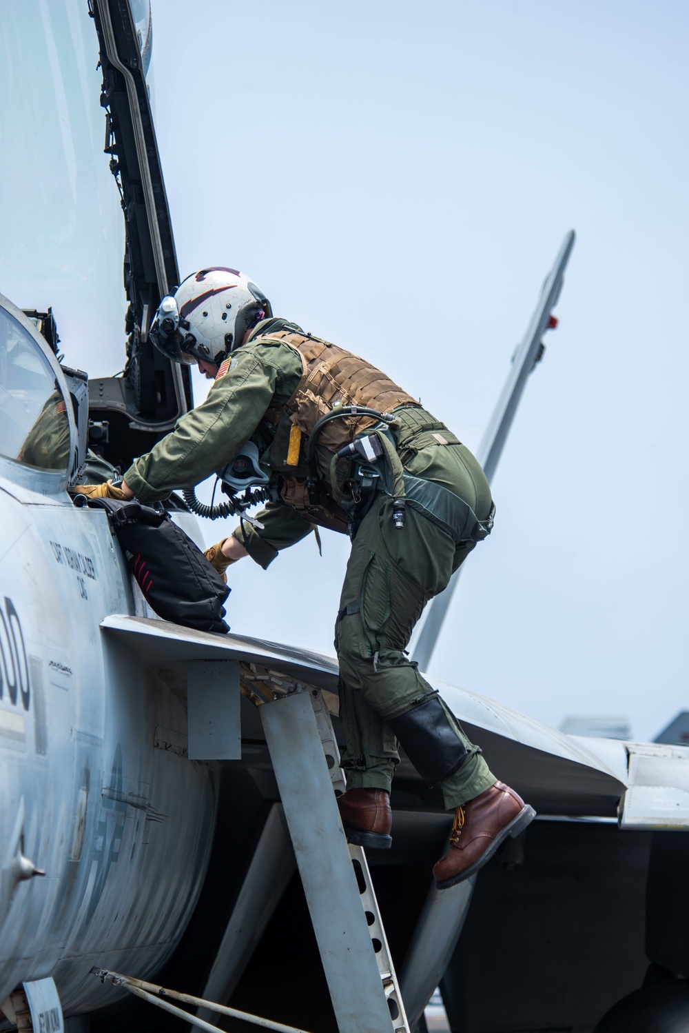 A military pilot in full flight gear is climbing into the cockpit of a jet aircraft, likely an F/A-18 Hornet or Super Hornet, using a boarding ladder. The pilot is wearing a green flight suit, brown tactical vest, white flight helmet with visor, and brown leather boots. The image captures a pre-flight boarding sequence on what appears to be a flight line or aircraft carrier deck.