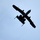 A military aircraft is photographed from below against a clear blue sky, appearing as a dark silhouette. The aircraft is banking at an angle, revealing its underside with twin engines mounted on the rear fuselage and straight wings. A faint contrail from a separate aircraft is visible crossing diagonally in the background.
