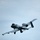 A military attack aircraft, identifiable as an A-10 Thunderbolt II (Warthog), is photographed in flight against an overcast light blue-gray sky. The aircraft is captured from a low angle, showing its underside with landing gear deployed, suggesting it may be on approach or shortly after takeoff. The aircraft is painted in standard gray camouflage livery.