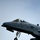 A close-up photograph of a U.S. Air Force A-10 Thunderbolt II (Warthog) attack aircraft, tail number 240, captured during takeoff or low-altitude flight with landing gear extended. The aircraft displays USAF markings including the distinctive star-and-bar insignia on the fuselage. A pilot is visible in the cockpit, and the aircraft's nose-mounted GAU-8 Avenger cannon barrel is clearly visible.