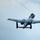 A military jet aircraft, identifiable as an A-10 Thunderbolt II (Warthog), is photographed from below at a low angle against an overcast sky. The aircraft is captured in flight, showing its distinctive twin-engine rear-mounted configuration and straight wings. The image appears to be taken at an airshow or during a low-altitude flyover.