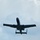 A military jet aircraft is photographed from below against an overcast sky, showing its underside in silhouette. The aircraft displays distinctive twin-engine rear-mounted configuration and twin tail booms, consistent with a close air support aircraft. The image is taken from a ground-level perspective looking upward.