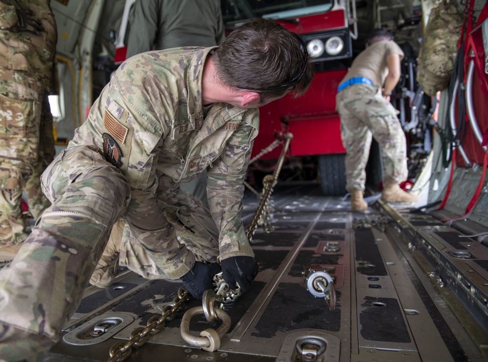 Military personnel in OCP (Operational Camouflage Pattern) uniforms are securing cargo chains to tie-down rings on the floor of a military transport aircraft. A large red vehicle, likely a fire truck or emergency vehicle, is visible in the background being loaded or secured. The scene depicts a standard military air cargo loading and restraint operation.