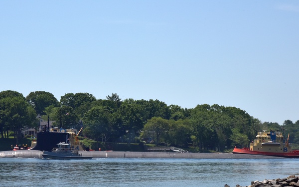 A large military submarine is transiting a river or harbor channel in surface mode, assisted by at least one tugboat. A prominent red and yellow tugboat marked with the number '8' is visible on the right side of the submarine, likely providing escort or maneuvering assistance. The scene is set against a tree-lined shoreline with residential structures visible in the background.