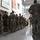 A large group of military personnel in camouflage uniforms are standing at parade rest inside the Mid-Ohio Foodbank facility. The soldiers appear to be assembled for a briefing or formation, facing toward a speaker or presenter at the far end of the room. The setting suggests a community service or humanitarian assistance mission at the food bank.