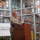 A U.S. Army soldier in camouflage uniform is speaking at a wooden podium during an event at the Mid-Ohio Food Collective facility. The setting appears to be inside a large warehouse or distribution center, with industrial shelving visible through glass windows in the background. The event appears to be an official address or announcement related to the food bank organization.