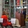 A formal press conference or official event is taking place inside what appears to be a food bank or warehouse facility. A man in a dark suit with a red tie is speaking at a wooden podium, with an American flag positioned behind him. Several attendees, including a person in military uniform and another in an orange outfit, are seated in reserved chairs facing the speaker.