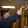 A man wearing a dark navy uniform and a black baseball cap with an American flag patch is reaching upward to work on or install equipment near the ceiling. He appears to be in an indoor facility with lockers visible in the background, suggesting a workplace or institutional setting. The man is handling what appears to be a flat panel or document near a white cabinet or equipment housing.