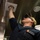 A military or naval personnel member wearing a dark blue uniform and tan cap is reaching up to inspect or adjust a ceiling-mounted fixture, likely a light or sensor. The setting appears to be an interior space with stainless steel surfaces, consistent with a ship's galley or utility area. The photo is taken from a low angle, looking upward at the subject.