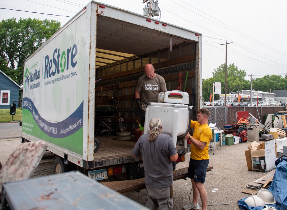 Three workers are loading or unloading a large white box truck belonging to Habitat for Humanity's ReStore program, a home improvement discount store. They are handling what appears to be a vintage or older-model washing machine at the rear of the truck. The scene takes place in a parking or staging area surrounded by various donated items and equipment.